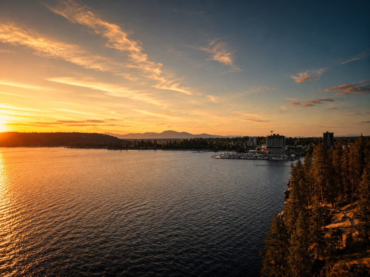 Golden sunset reflecting on a lake with a resort town, distant mountains, and pine trees.